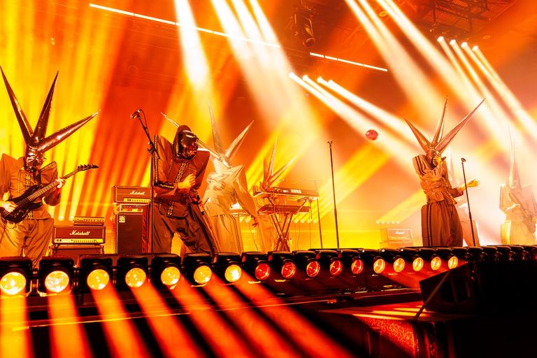Australian rock band TISM performing live on stage in masked costumes illuminated by bright yellow and orange spotlights.
